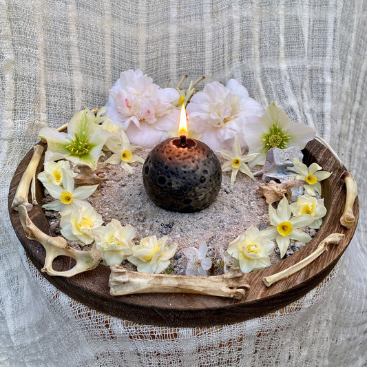 A black and white realistic Moon shaped beeswax candle on a wooden altar with ash, bones and early Spring flowers