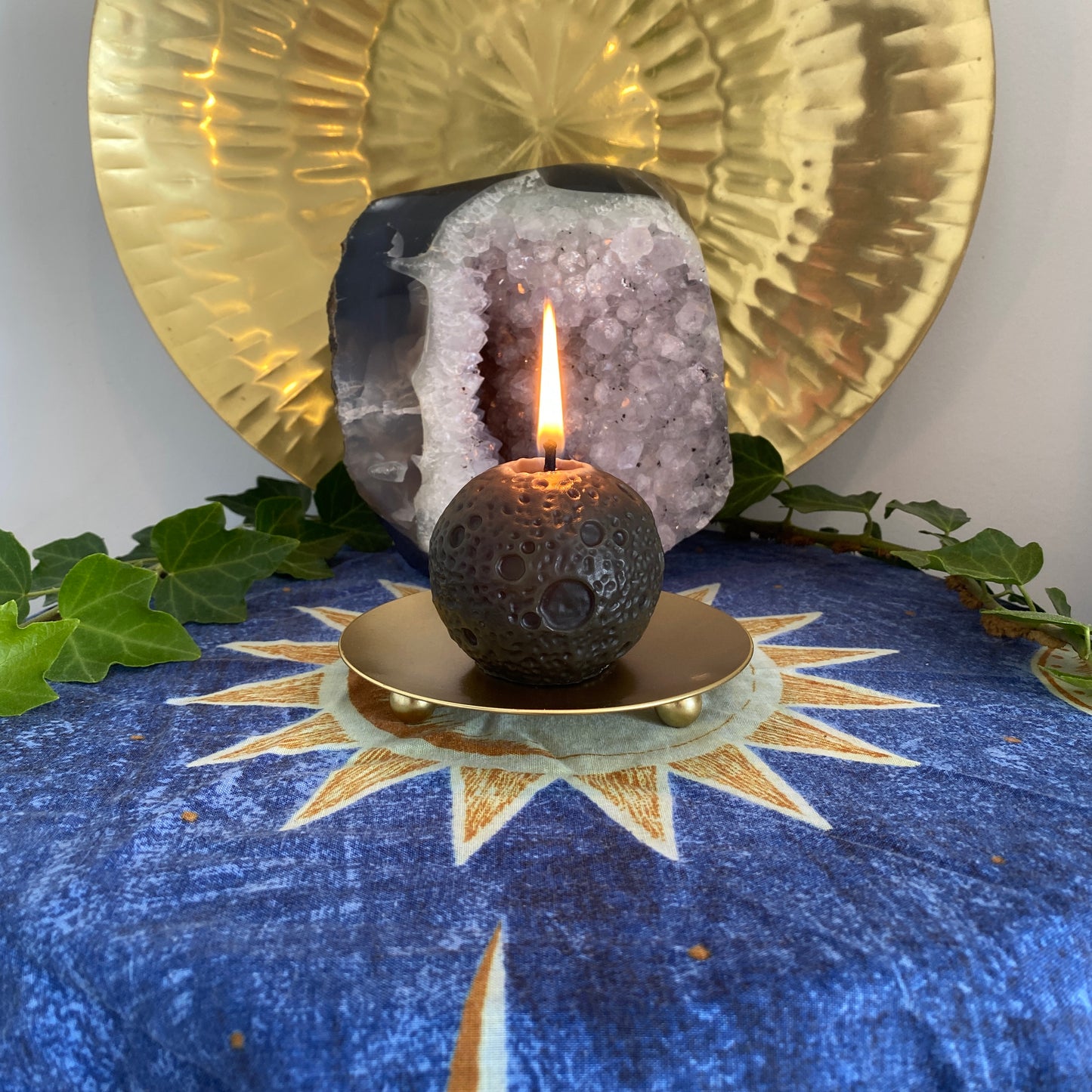 Decorative New Moon black beeswax candle with a lit flame on a blue cloth with Sun design, surrounded by gold plates, a large amethyst crystal and ivy.