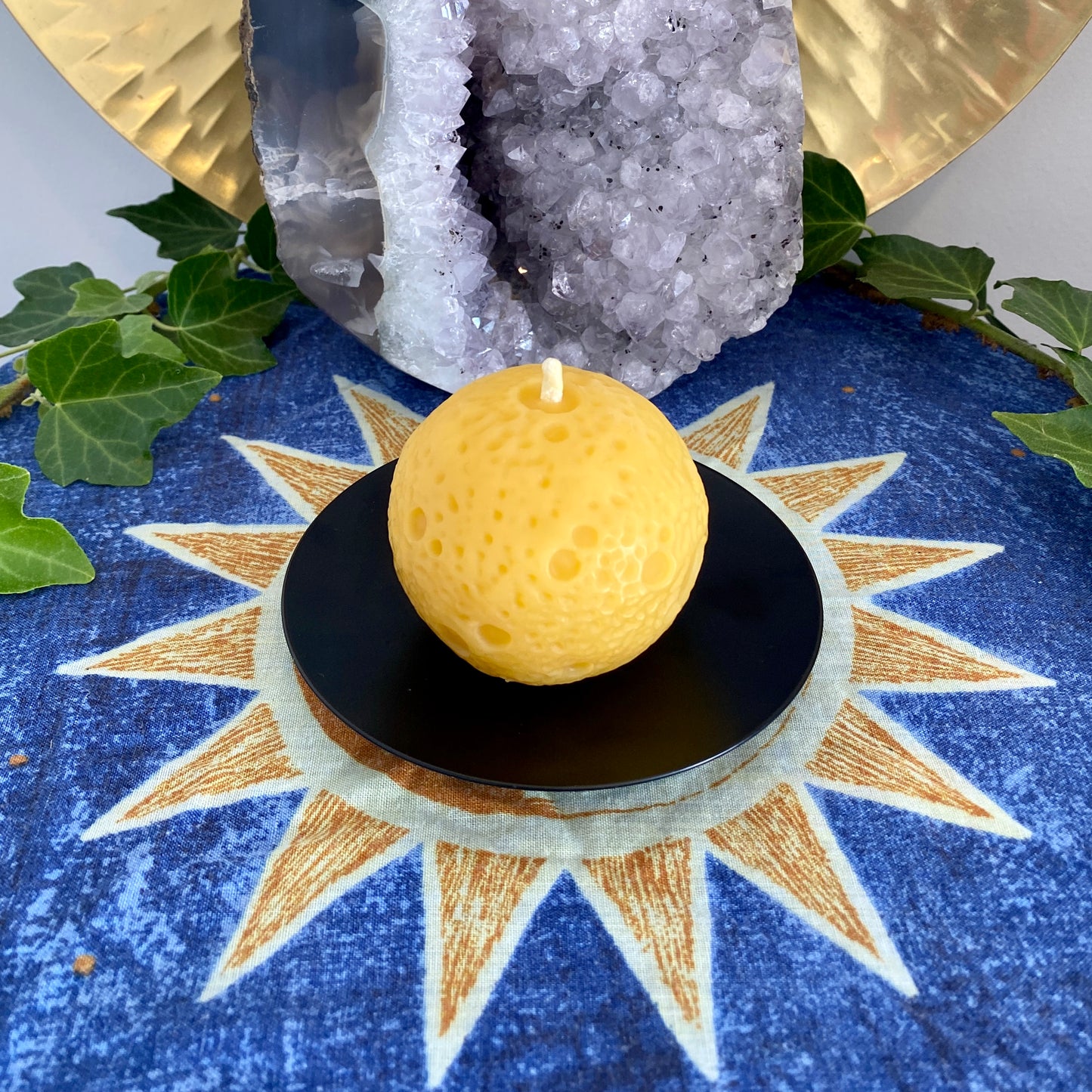 A realistic Moon-shaped golden beeswax candle displayed on a black holder with a sun and moon pattern cloth, accompanied by green leaves and a crystal in the background.