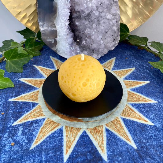 A realistic Moon-shaped golden beeswax candle displayed on a black holder with a sun and moon pattern cloth, accompanied by green leaves and a crystal in the background.