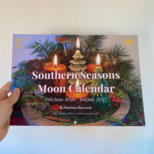 A hand holds a square printed calendar cover titled "Southern Seasons Moon Calendar" with a festive Yuletide scene showing lit candles, pine branches, pinecones, and a Sun and Moon. The cover text includes dates 15th June 2026 to 3rd July 2027 and author name By Maniana Heywood.