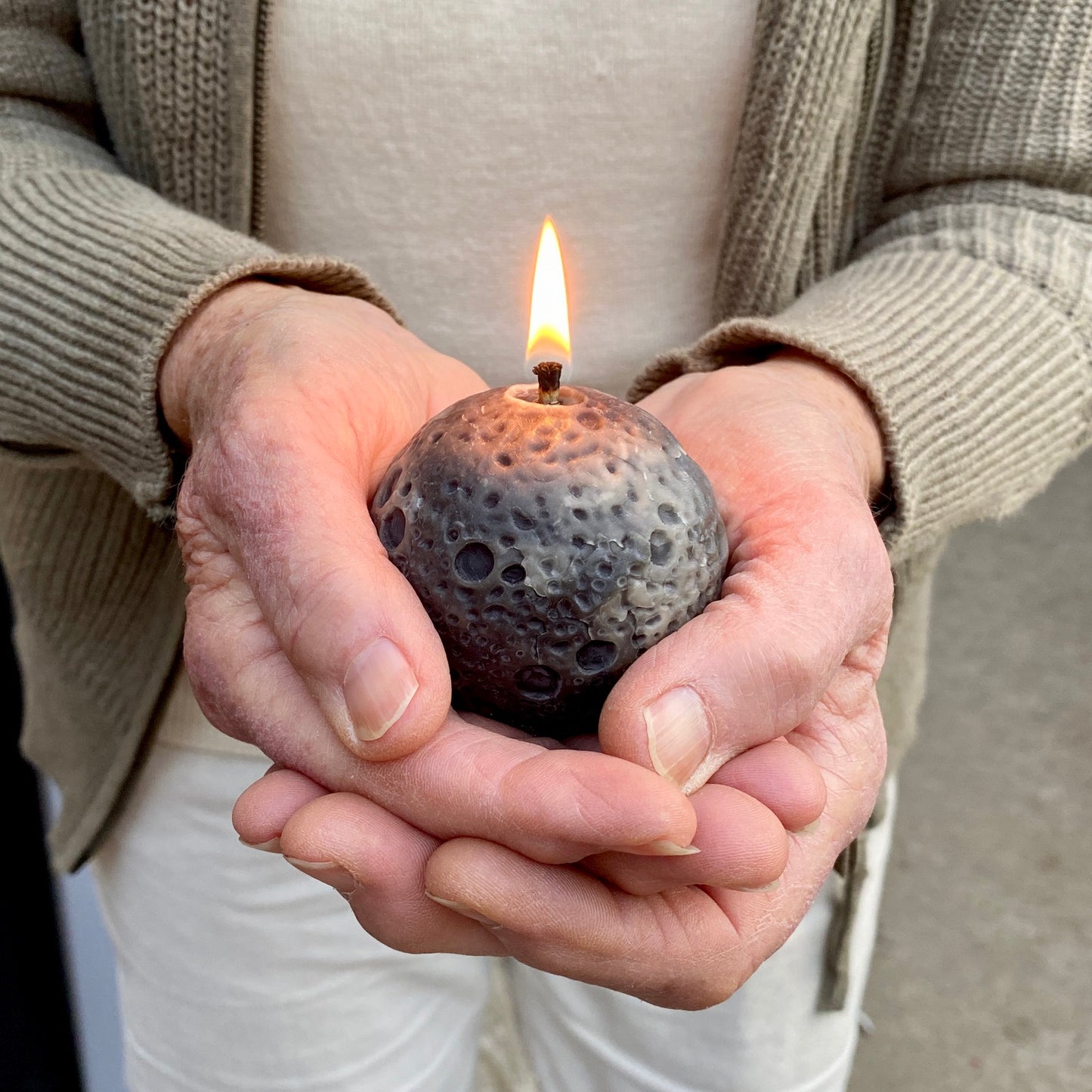 Older woman's hands holding a grey textured realistic Moon shaped candle with a lit flame. The person is wearing a beige cardigan and white pants.