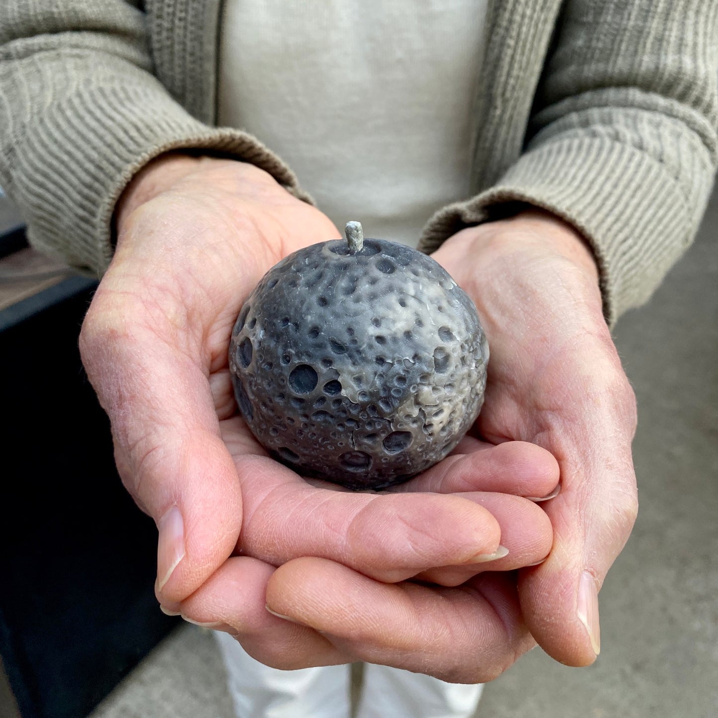 Older woman's hands holding a grey textured realistic Moon shaped candle. The person is wearing a beige cardigan and white pants.