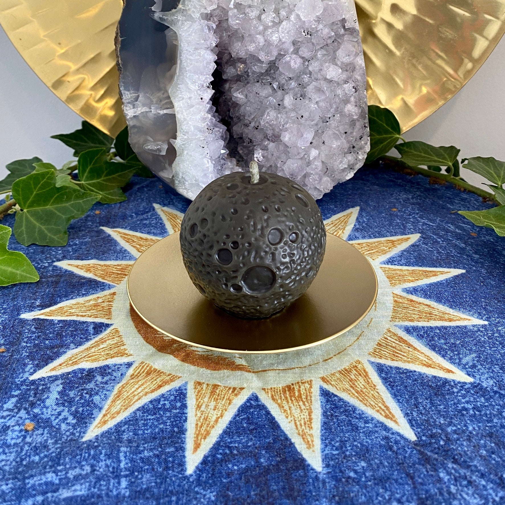 A realistic black moon-shaped beeswax candle displayed on a gold holder with a sun and moon pattern, accompanied by green leaves and a crystal in the background.