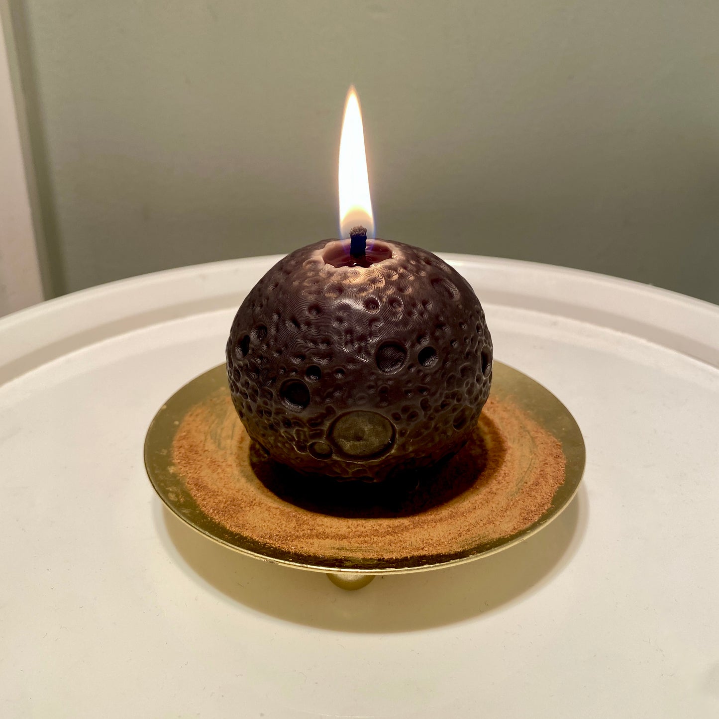 A realistic black moon-shaped beeswax candle with lit flame displayed on a gold holder with cinnamon powder on it and a white background.