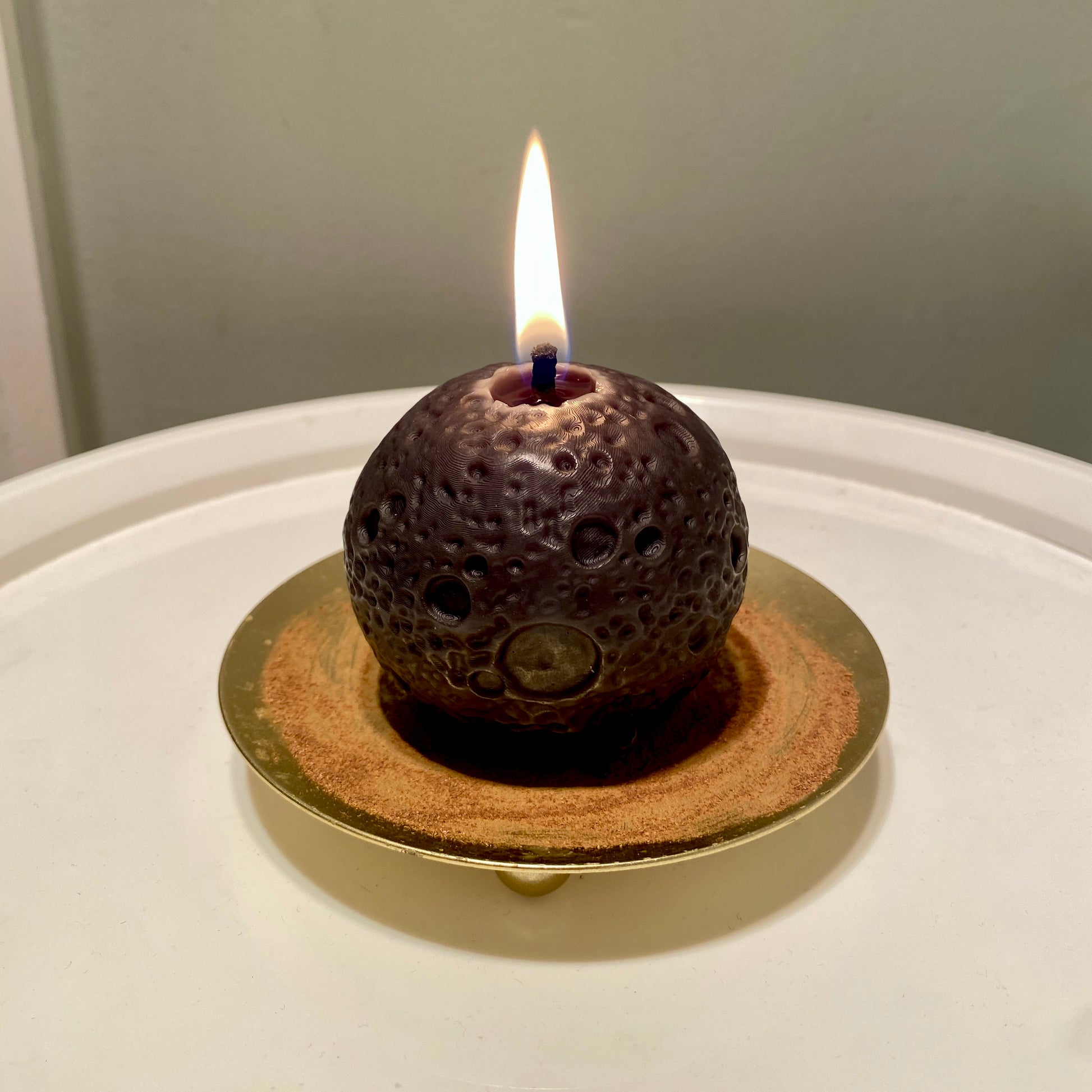A realistic black moon-shaped beeswax candle with lit flame displayed on a gold holder with cinnamon powder on it and a white background.