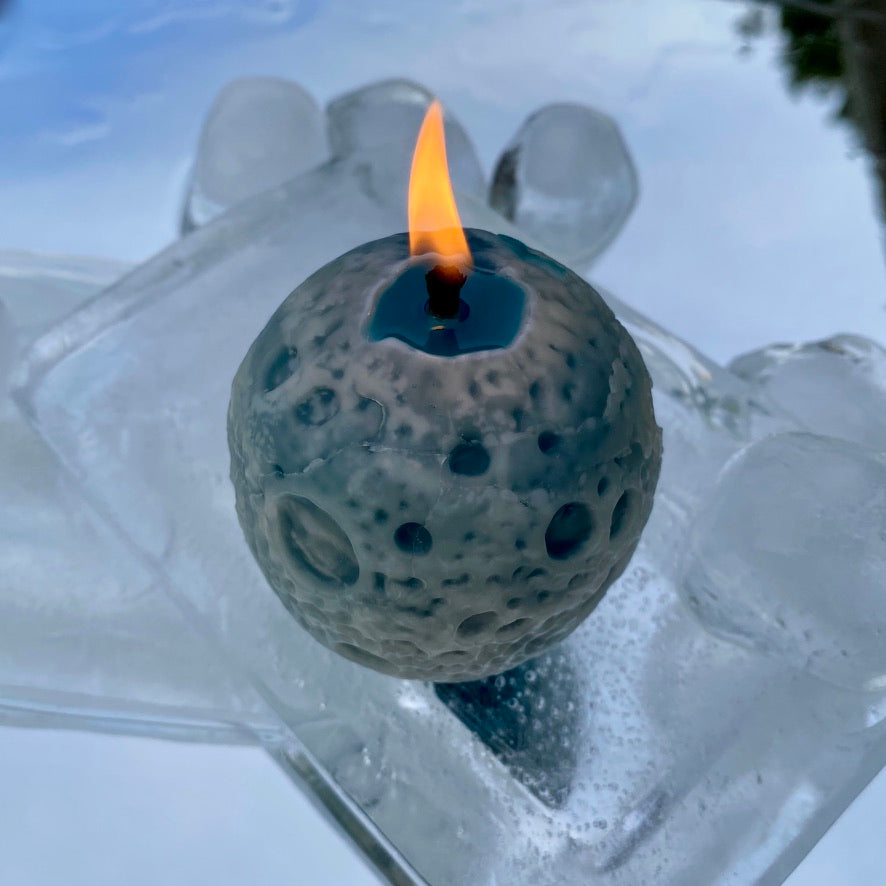 Spherical blue and white beeswax candle with textured surface and a burning flame that looks like a realistic Moon on an icy blue and white mirror background.