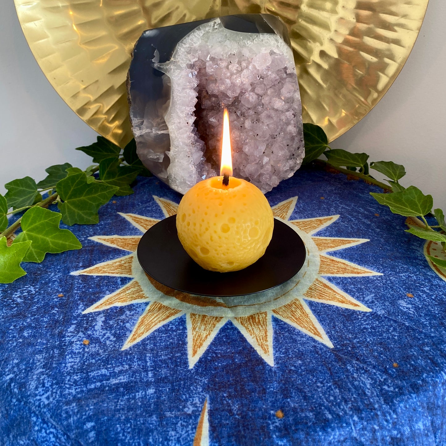 A realistic Moon-shaped golden beeswax candle with lit flame displayed on a black holder with a sun and moon pattern cloth, accompanied by green leaves and a crystal in the background.